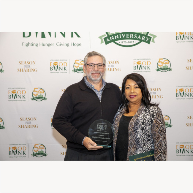 A man and woman stand together holding an award at a Food Bank event. The backdrop showcases the Los Angeles Regional Food Bank logo alongside the 50th-anniversary celebration and the text "Season for Sharing.