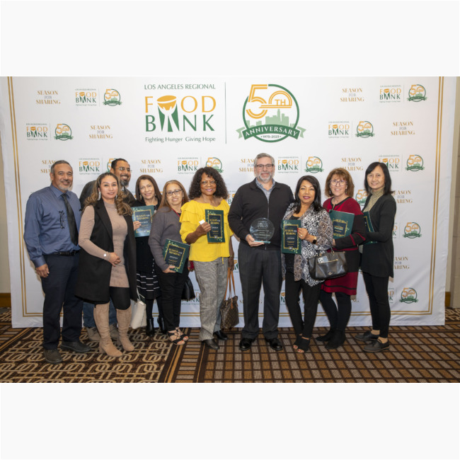 A group of people stands together, smiling, while holding plaques and an award in front of a backdrop celebrating the 50th anniversary of the Los Angeles Regional Food Bank.