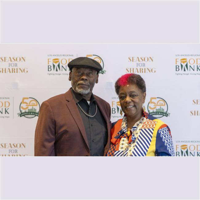 A man and woman are standing together, smiling in front of a "Los Angeles Regional Food Bank Season for Sharing 50th Anniversary" backdrop. The man sports a hat and blazer, while the woman dazzles in a colorful patterned outfit, embodying the festive spirit of the event.