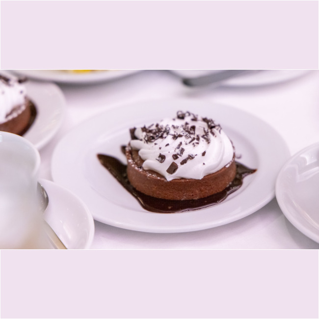 A chocolate dessert topped with whipped cream and chocolate shavings sits on a white plate, surrounded by a drizzle of chocolate sauce. It's placed on a table with partially visible dishes in the background, reminiscent of an elegant spread at a Los Angeles Regional Food Bank event.