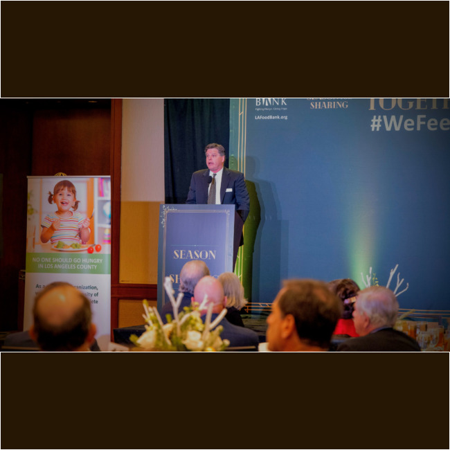 A man in a suit speaks at a podium labeled "Season of Sharing" at a formal event for the Los Angeles Regional Food Bank. Nearby, a sign depicts a child holding a bowl of food, while attendees are seated in the foreground, highlighting the community's commitment to ending hunger.