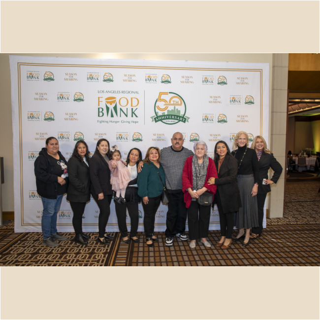 A group of people stands together, smiling in front of a Los Angeles Regional Food Bank backdrop, which prominently features their logo and a 50th-anniversary emblem. The setting is formal, highlighting the organization's commitment to fighting hunger in the community.