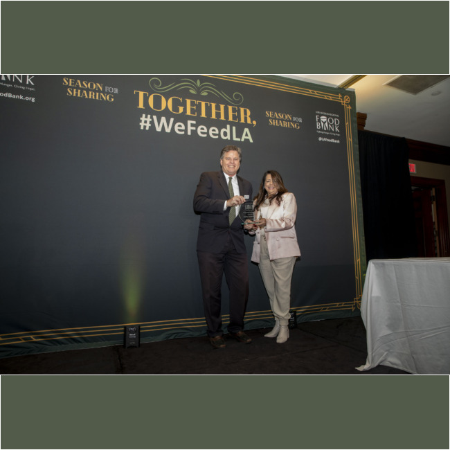 Two people stand on stage, proudly holding a glass award in front of a backdrop that reads "TOGETHER, Season for Sharing, #WeFeedLA," celebrating the efforts of the Los Angeles Regional Food Bank. They are dressed in formal attire.