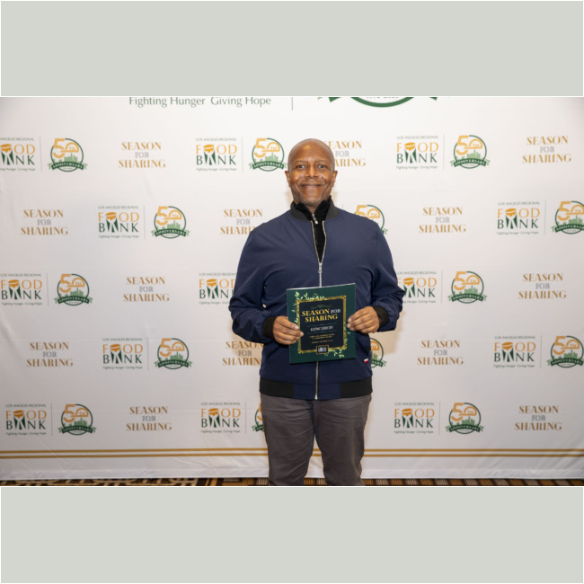 A person stands smiling, holding a "Season for Sharing" booklet in front of a backdrop adorned with "Food Bank" and "Season for Sharing" logos. They are wearing a dark jacket and grey pants, representing the spirit of giving at the Los Angeles Regional Food Bank.