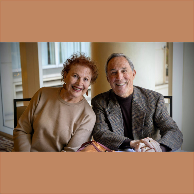 An elderly couple smiles warmly at the camera while sitting indoors, embodying the spirit of community supported by the Los Angeles Regional Food Bank. The woman wears a beige top and hoop earrings, while the man dons a checked blazer with a dark turtleneck. A window with blinds is seen in the background.