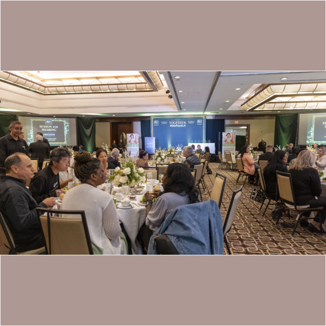 A large indoor event with round tables filled with people dining supports the Los Angeles Regional Food Bank. The stage at the front displays a "Together" banner. Warm lighting and floral centerpieces adorn each table as attendees engage in lively conversation, united for a cause.