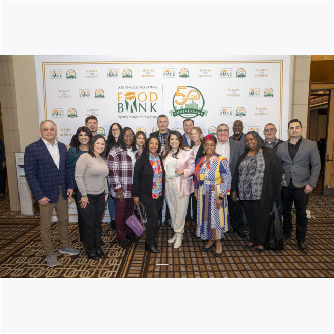 A diverse group of people stands together, beaming in front of a backdrop adorned with the Los Angeles Regional Food Bank's logo and 50th Anniversary design. Dressed in both business and casual attire, their smiles reflect a shared commitment to fighting hunger in the community.