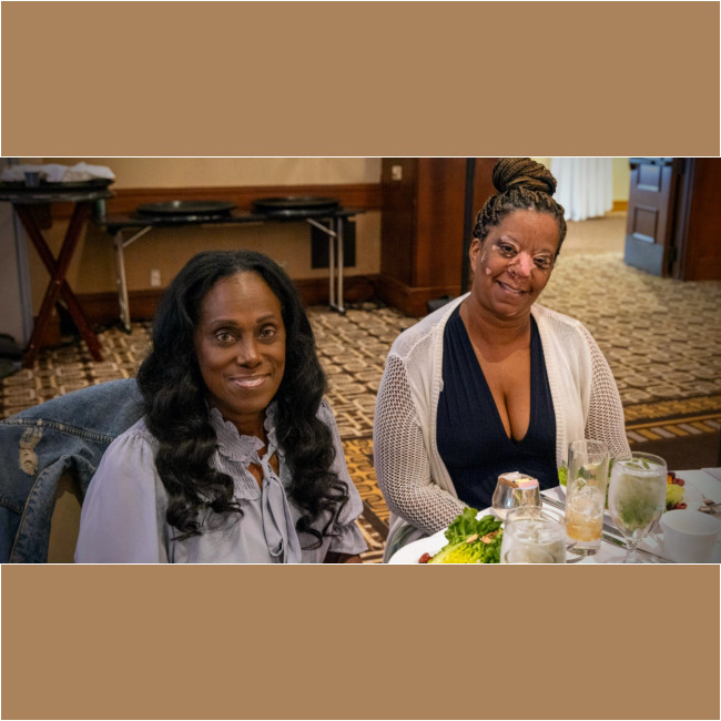 Two women are sitting at a table in a restaurant, enjoying a meal that supports the Los Angeles Regional Food Bank. The table is adorned with plates of food, drinks, and a colorful dish. One wears a light blouse while the other sports a white cardigan over a dark top, both smiling warmly at the camera.