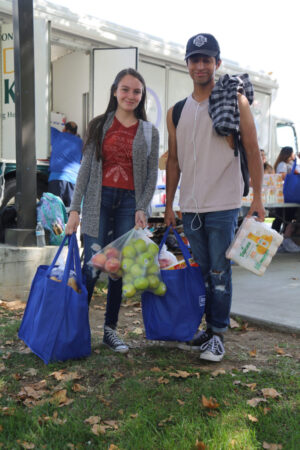 Two people stand outdoors holding reusable bags filled with groceries, including apples, smiling as a Los Angeles Regional Food Bank food truck is visible in the background. A few autumn leaves are scattered on the grass around them.