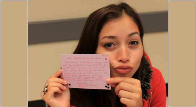 Writing letters filled with gratitude and appreciation to the families of active service members. A young woman in red holds up a written index card addressed to families of active service expressing gratitude.