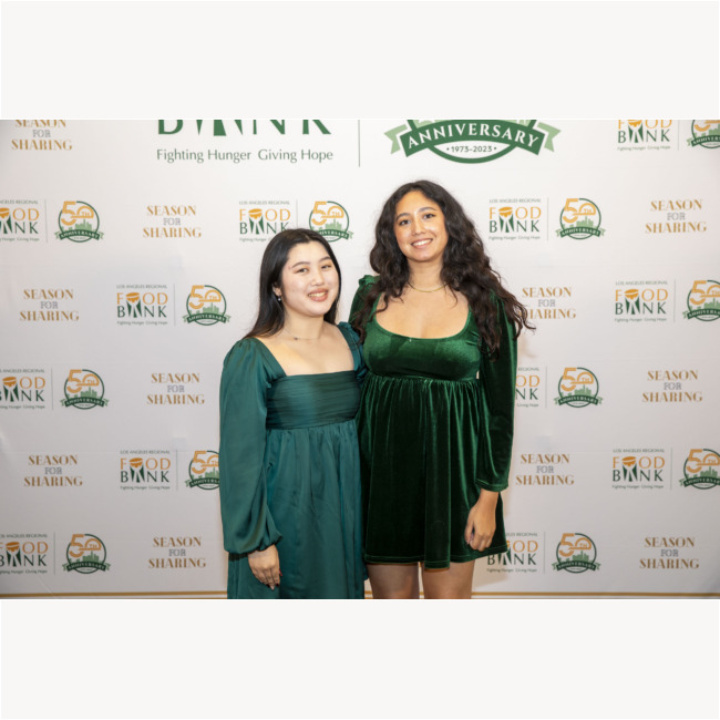 Two women in green dresses stand smiling in front of a backdrop featuring logos for the Los Angeles Regional Food Bank, "Season for Sharing," and an anniversary message. The backdrop is adorned with vibrant green and orange accents.
