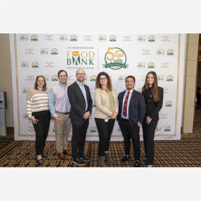 A group of six people, dressed in business attire, is smiling for the camera in front of a backdrop adorned with logos celebrating the Los Angeles Regional Food Bank's 50th anniversary.