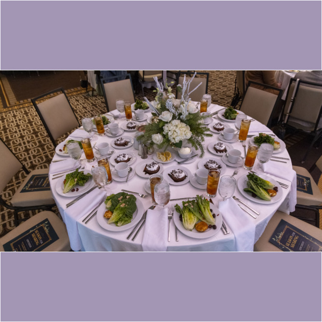 A round dining table set with white linens, elegantly adorned with a floral centerpiece, evokes the spirit of the Los Angeles Regional Food Bank. The table boasts plates with salads, dessert dishes, iced tea, water, and cutlery. Each seat has a menu placed on the chair.