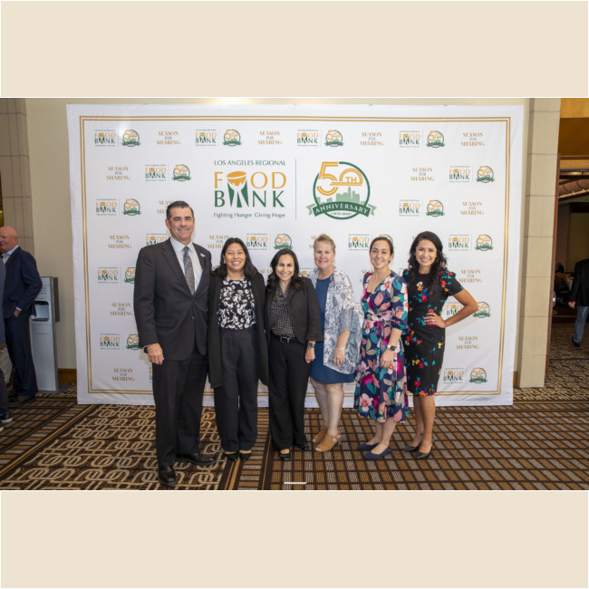A group of six stands smiling in front of a Los Angeles Regional Food Bank's 50th anniversary banner. Dressed in business and casual attire, they pose on a patterned carpet, celebrating the impact and legacy of the food bank in their community.
