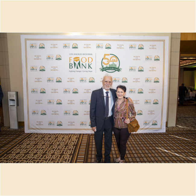 An older man in a suit and an older woman in casual attire stand smiling together in front of a backdrop featuring the Los Angeles Regional Food Bank 50th Anniversary logos, capturing the joyful spirit of this meaningful event.