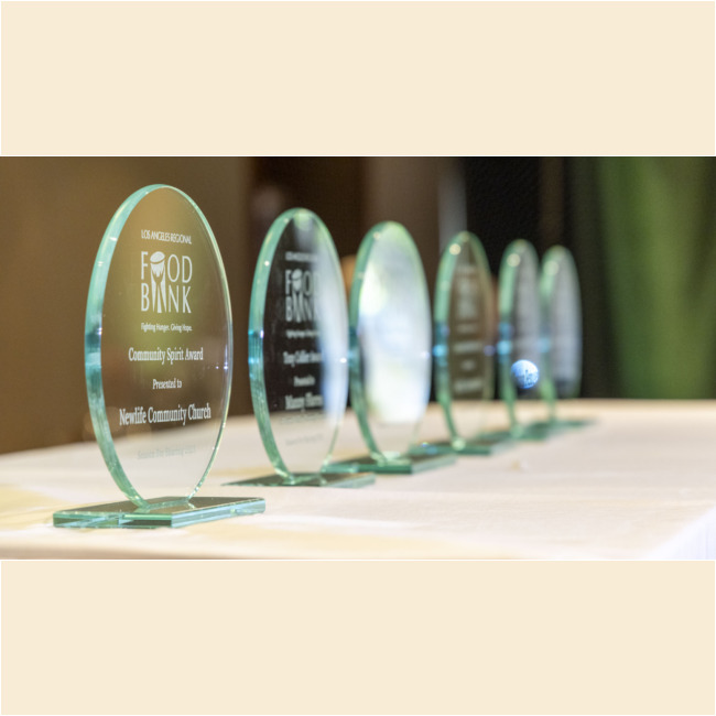 Several glass awards are lined up on a white table, each inscribed with "Food Bank Community Spirit Award" and reflecting light. The closest award honors "NewLife Community Church" for its efforts in collaboration with the Los Angeles Regional Food Bank.