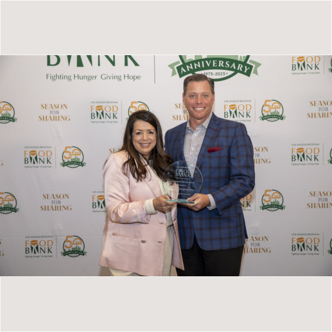Two people stand together smiling, proudly holding an award at the Los Angeles Regional Food Bank event. The backdrop showcases logos and text celebrating the Los Angeles Regional Food Bank's anniversary and mission.
