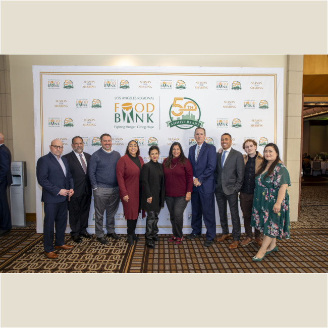 A group of people stands in front of a backdrop with the Los Angeles Regional Food Bank logo, joyfully celebrating its 50th anniversary. Dressed in both business and casual attire, they smile warmly at the camera, embodying a shared commitment to community support.