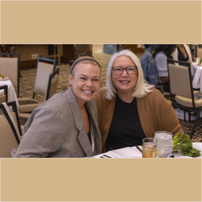 Two smiling women seated at a dining table, one with short, blonde hair and a plaid blazer, the other with long, blonde hair and glasses. Plates, drinks, and a salad from the Los Angeles Regional Food Bank grace the table in a warmly lit room.