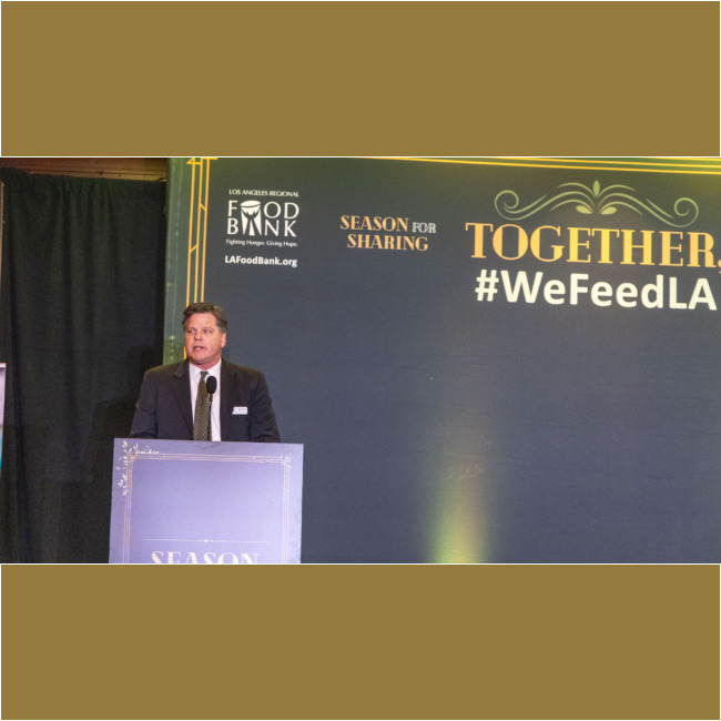 A man in a suit stands at a podium, passionately addressing an event for the Los Angeles Regional Food Bank, with "Los Angeles Regional Food Bank" prominently displayed and hashtags like "#WeFeedLA" and "Season for Sharing." The formal setting features elegant decorative elements that enhance the occasion.
