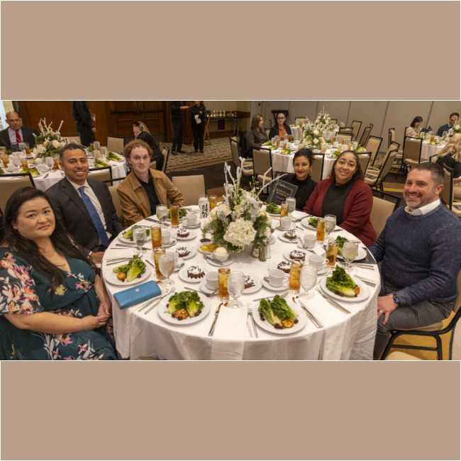 A group of people sit around a round banquet table set with salads, desserts, and drinks. The table features a floral centerpiece. Other guests and tables are visible in the background at this Los Angeles Regional Food Bank event.