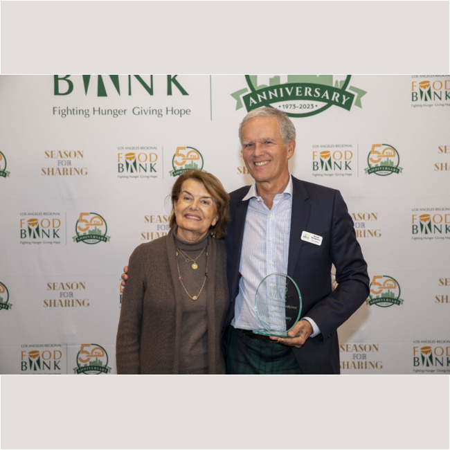 A man and a woman stand smiling together in front of a backdrop celebrating the Los Angeles Regional Food Bank's anniversary. The man proudly holds a glass award, showcasing their commitment to fighting hunger.