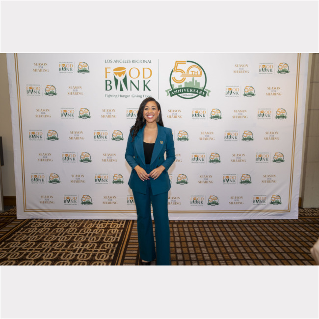 A woman in a blue suit stands smiling on a carpeted floor, framed by the Los Angeles Regional Food Bank logo and a 50th-anniversary emblem.