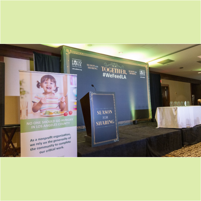 A conference room features a podium displaying "Season for Sharing." Nearby, a banner with a smiling child declares, "No one should go hungry in Los Angeles County." Awards are arranged on a table, while the backdrop proudly highlights "#WeFeedLA" and "Together" alongside the Los Angeles Regional Food Bank.