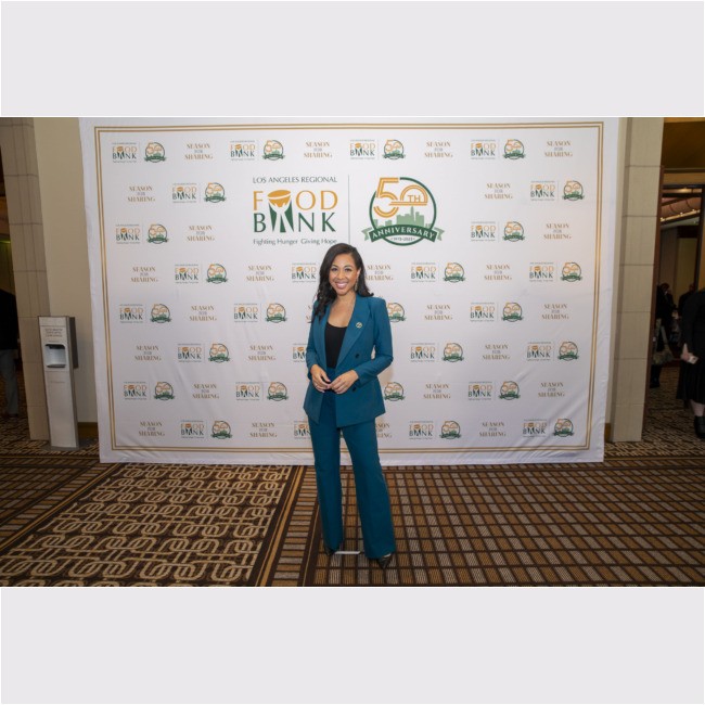 A woman in a turquoise suit poses gracefully before the Los Angeles Regional Food Bank's 50th Anniversary backdrop, adorned with its emblematic logos and celebratory text.