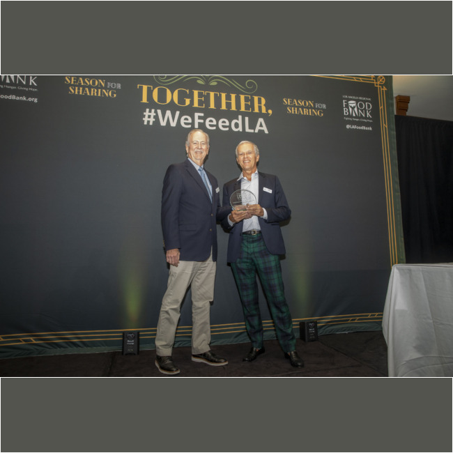 Two men stand smiling in front of a "#WeFeedLA" backdrop, proudly holding an award. Clad in suits, with one sporting plaid pants, they celebrate the success of the "Season for Sharing" initiative by the Los Angeles Regional Food Bank.