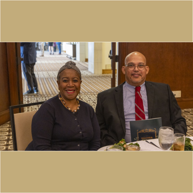 A man in a suit and tie and a woman in a black top sit at a table with plates of food and drinks, smiling at the camera. The warmly lit dining area, featuring patterned carpet, is part of an event supporting the Los Angeles Regional Food Bank.