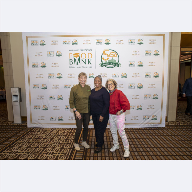 Three people stand smiling in front of a backdrop adorned with the Los Angeles Regional Food Bank's 50th Anniversary logo. The carpeted floor complements the background, which features repeating logos and celebratory text for this milestone occasion.