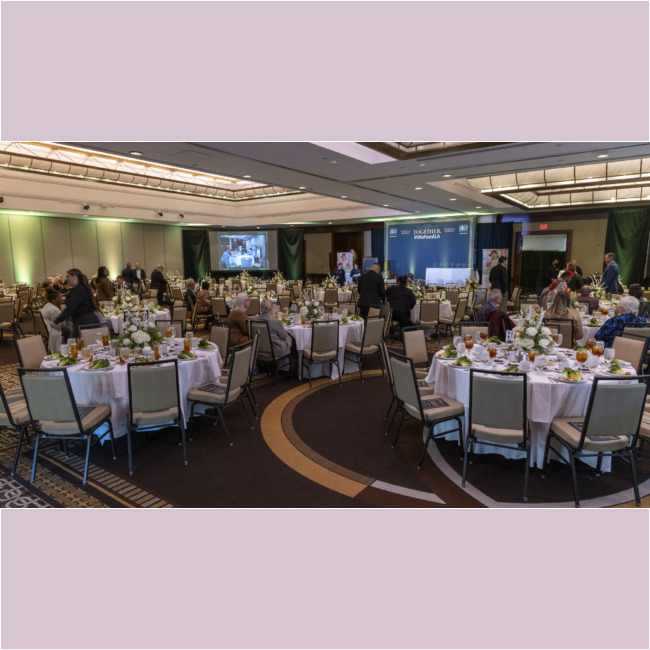 A large banquet hall set for a formal event hosted by the Los Angeles Regional Food Bank. Round tables with white tablecloths boast floral centerpieces, tableware, and glasses. Guests are seated and mingling as a stage and screen loom in the background.