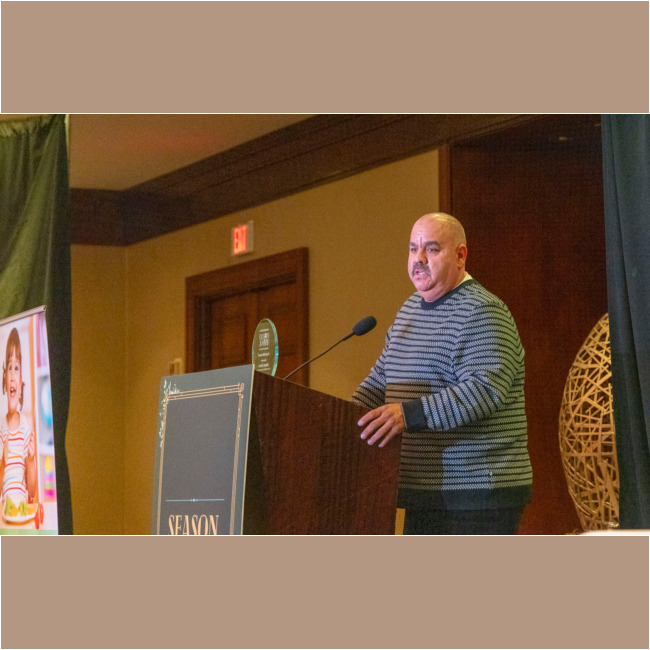 A man in a striped sweater stands at a podium adorned with decorative gold lettering, speaking into a microphone. Nearby, a poster of a smiling child partly catches the eye, while the backdrop showcases a large decorative ball. The event is supported by the Los Angeles Regional Food Bank.