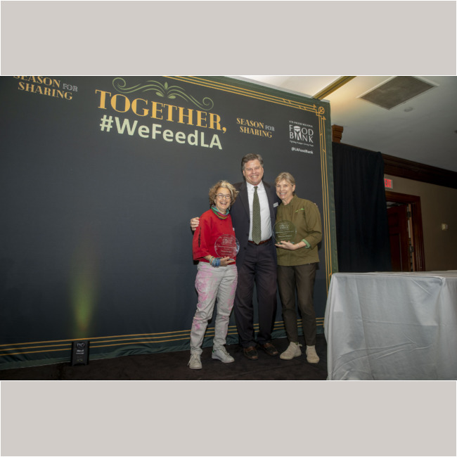 Three people stand on stage holding awards in front of a backdrop with "Together, #WeFeedLA." They smile at the camera, dressed in formal and casual attire. The event is organized by the Los Angeles Regional Food Bank.