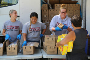 Four people stand by an open truck, distributing packaged food. Three wear matching "Citrus Water Polo" shirts and blue gloves. They smile while handing items to a woman with short hair holding a box. Stacks of Los Angeles Regional Food Bank boxes fill the truck in the background.