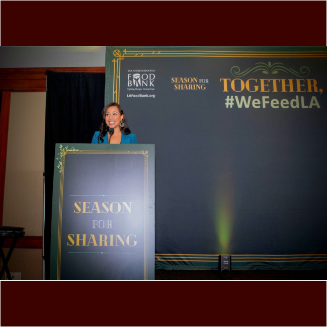 A woman stands at a podium labeled "Season for Sharing" with the Los Angeles Food Bank logo, part of the Los Angeles Regional Food Bank's initiative. She addresses an audience against a backdrop reading "TOGETHER, #WeFeedLA," highlighting community efforts to fight hunger.