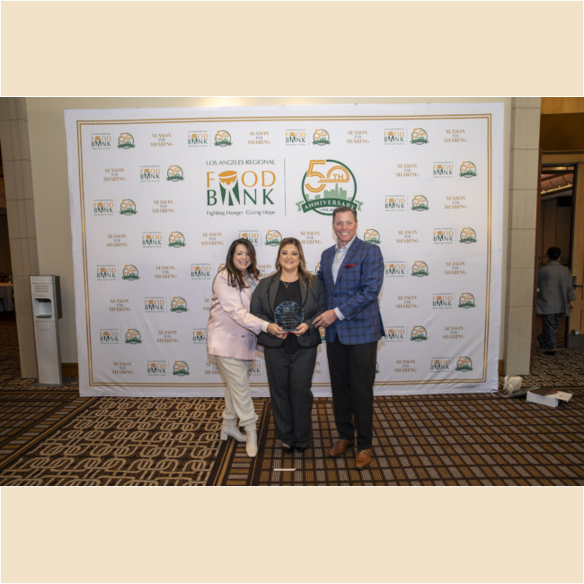 Three people stand together smiling, proudly holding an award in front of a Los Angeles Regional Food Bank 50th Anniversary backdrop. The woman in the center wears black, the person on the left is in pink, and the person on the right sports a blue plaid jacket.