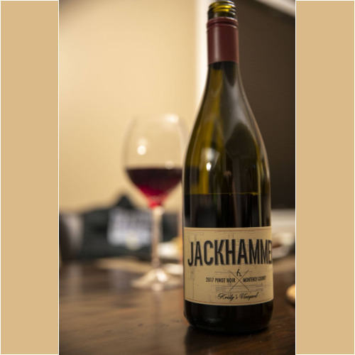 A bottle of Jackhammer 2017 Pinot Noir from Monterey County graces the wooden table, with a filled wine glass subtly suggesting indulgence. In the blurred indoor setting, it quietly echoes gatherings supporting causes like the Los Angeles Regional Food Bank.