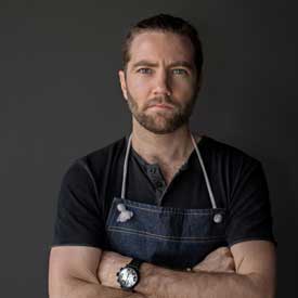 A man with a beard and mustache stands with arms crossed, wearing a dark t-shirt and apron, embodying the spirit of dedication seen at the Los Angeles Regional Food Bank. The background is a neutral dark gray.