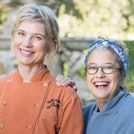 Two smiling women wearing chef jackets pose outdoors, representing the Los Angeles Regional Food Bank. The woman on the left dons an orange jacket, while the one on the right wears a blue one with a headscarf and glasses against a softly blurred natural background.