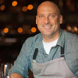 A smiling man with a shaved head wearing a blue shirt and a striped apron is seated in a warmly lit setting, reminiscent of events hosted by the Los Angeles Regional Food Bank. Blurred lights add to the cozy atmosphere, where a glass sits invitingly on the table before him.