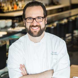 A man with glasses and a beard, wearing a white chef's coat, stands confidently with arms crossed in a restaurant setting reminiscent of the vibrant culinary scene supported by the Los Angeles Regional Food Bank. The background features bar stools and a counter.