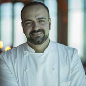 A man with a beard and shaved head is smiling in his white chef's coat. Indoors, under soft lighting, he stands as a proud supporter of the Los Angeles Regional Food Bank, embodying his commitment to community nourishment.