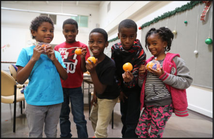 Five children stand in a room, smiling and holding oranges donated by the Los Angeles Regional Food Bank. They are casually dressed and appear to be enjoying themselves. The room is decorated with garlands and ornaments, creating a festive atmosphere.