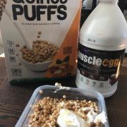 A container filled with quinoa puffs and yogurt sits on a table. Behind it, a box labeled "Quinoa Puffs" and a container marked "MuscleEgg" are neatly aligned, reminiscent of the variety you'll find at the Los Angeles Regional Food Bank. Quinoa puffs spill from the box into a bowl on the packaging.