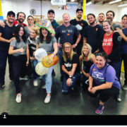 A group of people pose indoors, smiling at the camera. One person stands in front holding balloons and wearing a colorful hat. Everyone appears relaxed and happy, suggesting a celebratory occasion at the Los Angeles Regional Food Bank. The background features industrial elements.