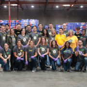 A group of people wearing casual clothes and gloves, posing indoors in a warehouse with stacks of boxes behind them. Some don matching T-shirts, suggesting they’re a team or volunteer group at the Los Angeles Regional Food Bank.