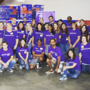 A group of people in matching purple shirts joyfully pose for a group photo indoors, embodying their dedication to the Los Angeles Regional Food Bank. Smiling faces beam as boxes are stacked in the background, symbolizing their united effort in tackling hunger.