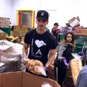 A person wearing a black cap and "I ❤️ LA" shirt is holding a bag of potatoes at the Los Angeles Regional Food Bank. In the background, another individual manages a loaf of bread, surrounded by stacks of boxes and packaged goods.